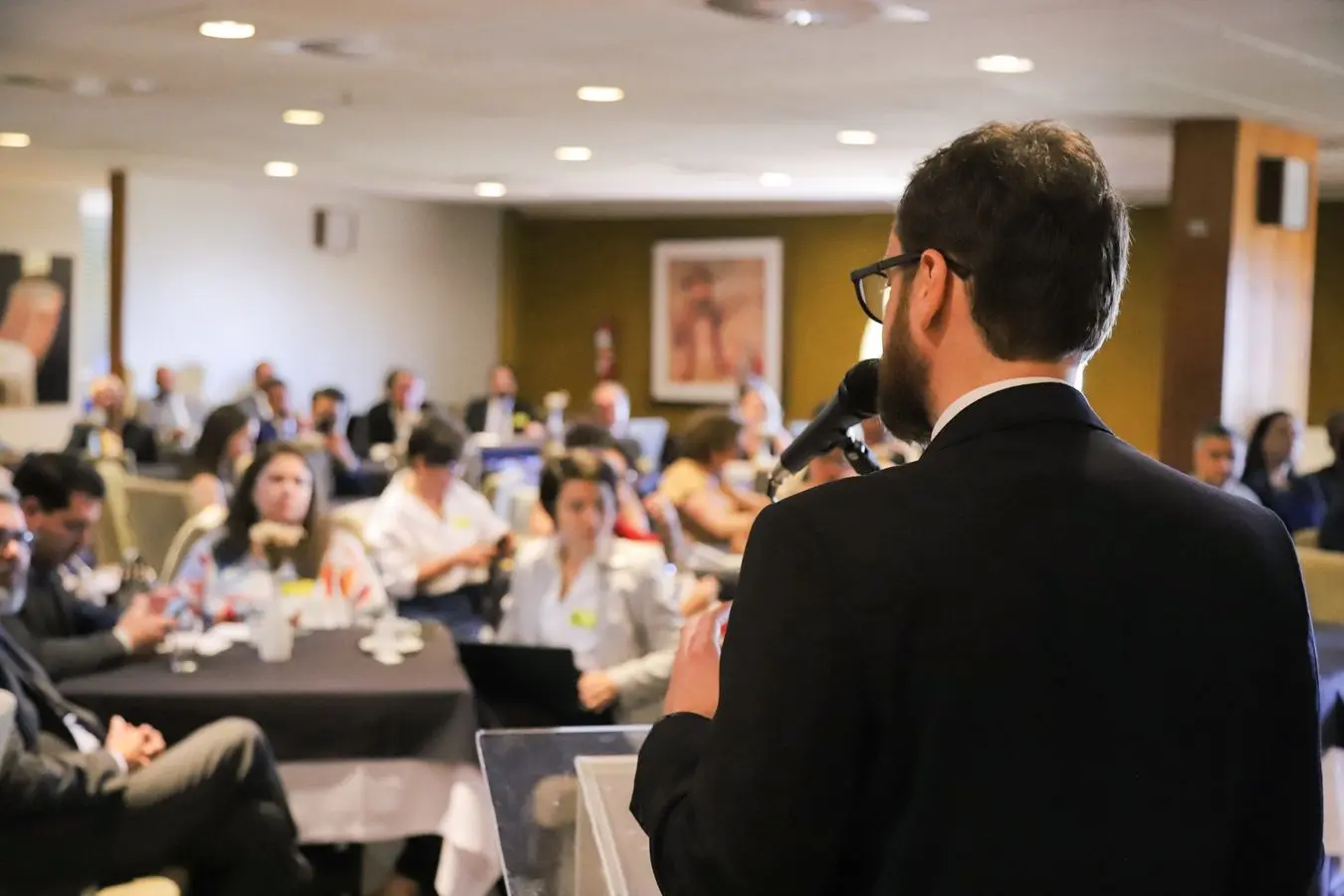 Rafael Arcuri, de costas, fala ao microfone para o público do Café da Manhã de Diálogos